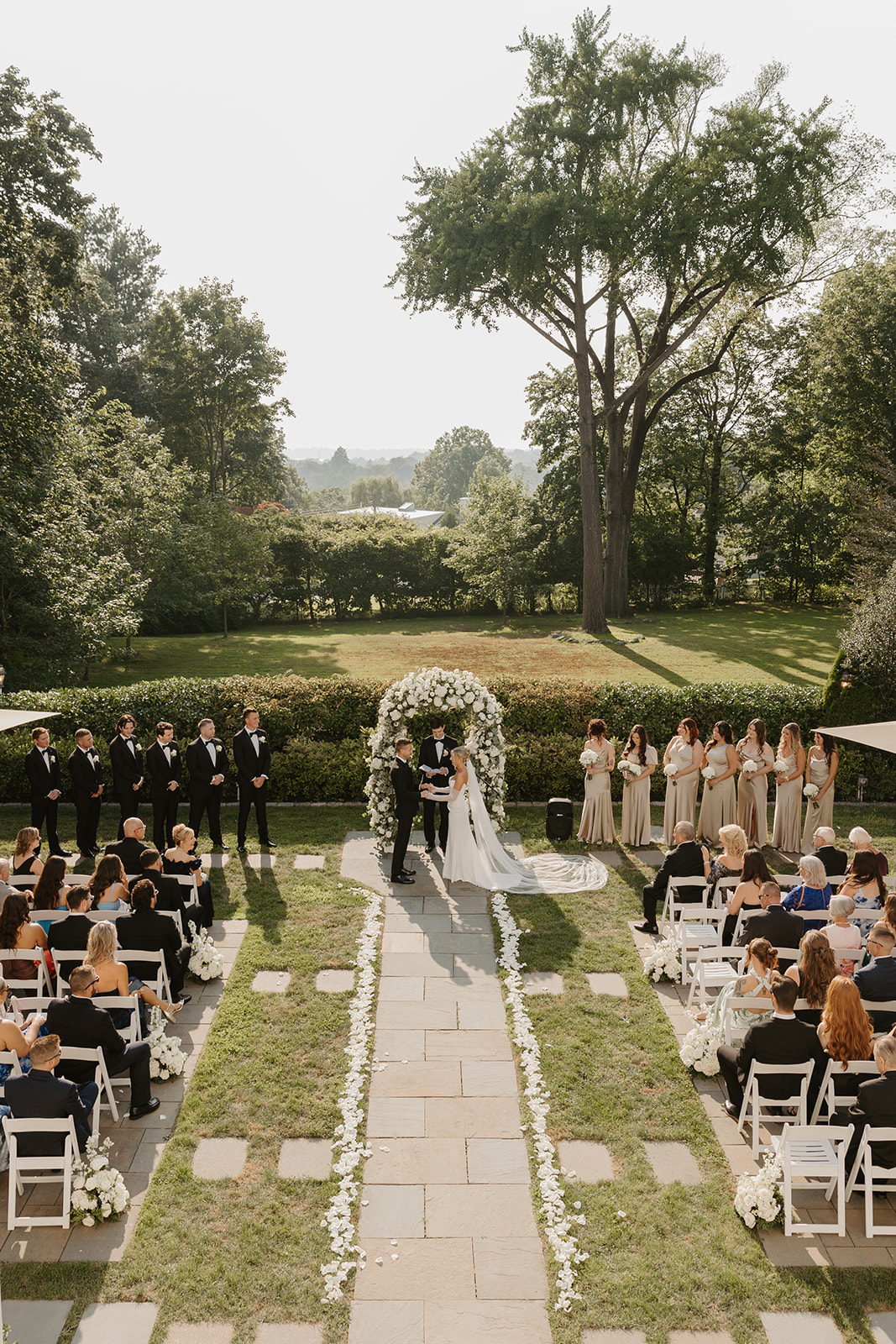 Wide high-angle view of the wedding ceremony under a blue sky, with the couple at the floral altar.